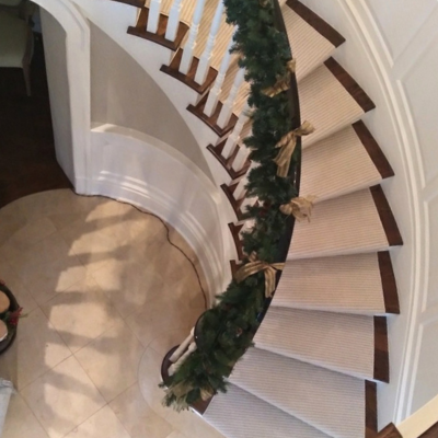 A curved staircase with a beige stair runner rugs and decorated with a garland of evergreen branches and gold bows for the holidays.